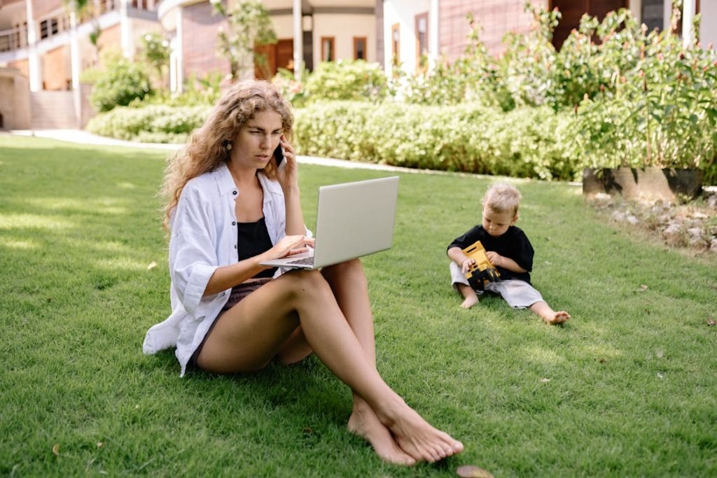 Woman Using Laptop While Sitting on Grass Field With Her Child